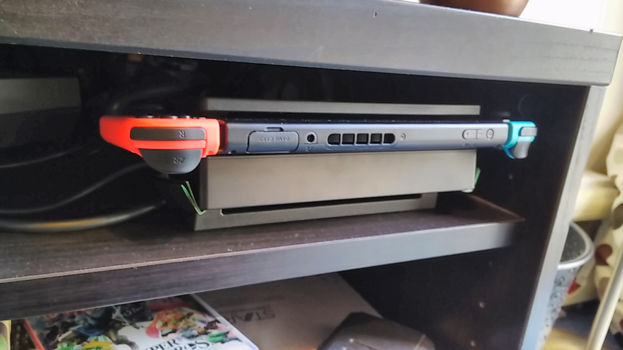 Nintendo switch in a dock, laying flat on a shelf, with the flap at the bottom slightly open.  The top of the switch is facing outwards